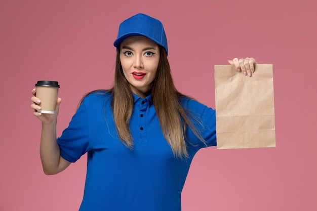 Mensajero femenino de vista frontal en uniforme azul y capa con paquete de comida de taza de café de entrega en la pared rosa