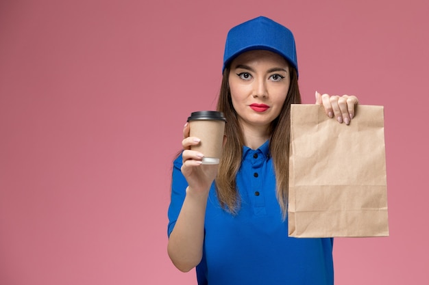 Mensajero femenino de vista frontal en uniforme azul y capa con paquete de comida de taza de café de entrega en la pared rosa