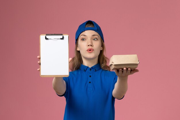 Mensajero femenino de vista frontal en uniforme azul y capa con pequeño paquete de comida de entrega y bloc de notas en el empleado del servicio de entrega de escritorio rosa