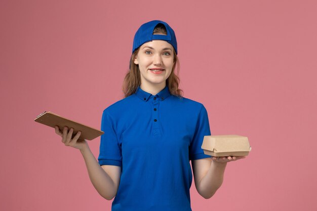 Mensajero femenino de vista frontal en uniforme azul y capa con pequeño paquete de comida de entrega y bloc de notas en la pared rosa, empleada del servicio de entrega de niña