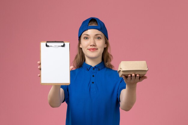 Mensajero femenino de vista frontal en uniforme azul y capa con pequeño paquete de comida de entrega y bloc de notas en la pared rosa, empleado del servicio de entrega