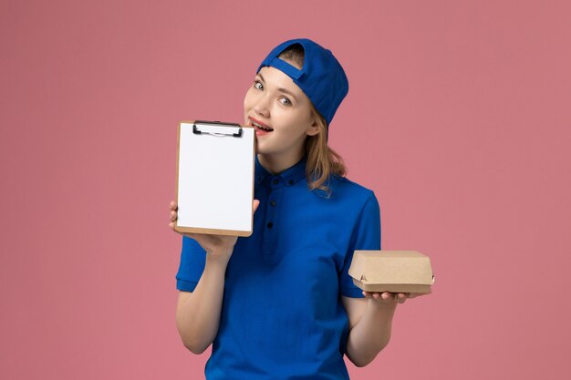 Mensajero femenino de vista frontal en uniforme azul y capa con pequeño paquete de comida de entrega y bloc de notas en la pared rosa, empleado del servicio de entrega