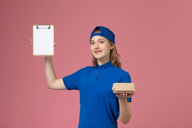 Mensajero femenino de vista frontal en uniforme azul y capa con pequeño paquete de comida de entrega y bloc de notas en la pared rosa, trabajo de empleado de servicio de entrega