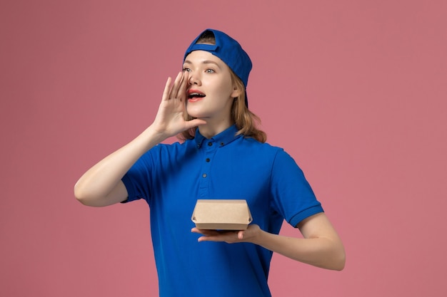 Mensajero femenino de vista frontal en uniforme azul y capa con pequeño paquete de comida de entrega en el fondo rosa empresa de servicio uniforme de entrega trabajo niña trabajo