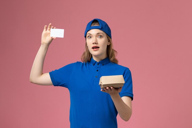 Mensajero femenino de vista frontal en uniforme azul y capa con pequeño paquete de comida de entrega con tarjeta en la pared rosa, trabajo de empleado de servicio de entrega