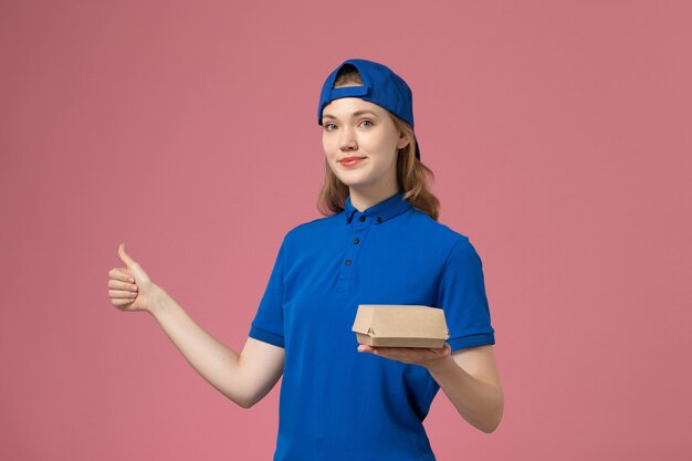 Mensajero femenino de vista frontal en uniforme azul y capa con pequeño paquete de comida de entrega en el trabajo de trabajo de empresa de servicio uniforme de entrega de fondo rosa