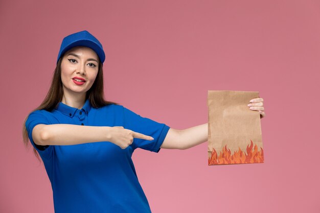 Mensajero femenino de vista frontal en uniforme azul y capa que sostiene el paquete de alimentos de papel en el trabajador de la pared rosa