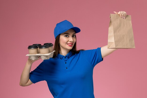 Mensajero femenino de la vista frontal en uniforme azul y capa que sostiene el paquete de comida de las tazas de café en la pared rosada