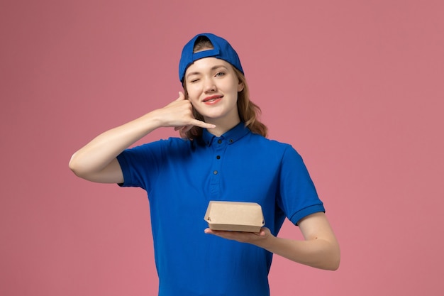 Mensajero femenino de la vista frontal en uniforme azul y capa que sostiene el pequeño paquete de comida de entrega en el trabajo de la muchacha del trabajador del trabajo de la empresa uniforme de entrega del fondo rosa