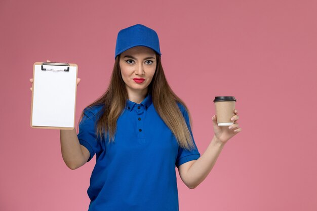 Mensajero femenino de vista frontal en uniforme azul y capa sosteniendo el bloc de notas de la taza de café de entrega en la pared rosa