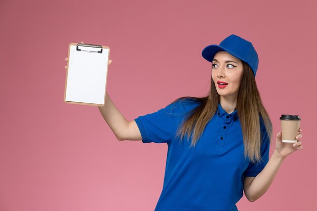 Mensajero femenino de vista frontal en uniforme azul y capa sosteniendo el bloc de notas de la taza de café de entrega en la pared rosa