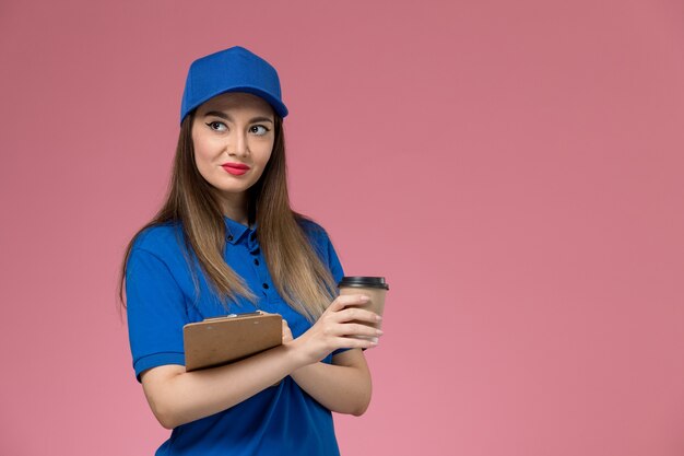 Mensajero femenino de vista frontal en uniforme azul y capa sosteniendo el bloc de notas de la taza de café de entrega en la pared rosa