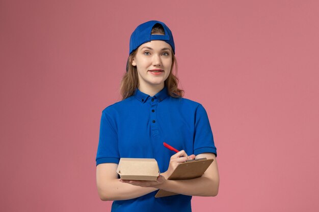 Mensajero femenino de vista frontal en uniforme azul y capa sosteniendo un pequeño bloc de notas de paquete de comida de entrega y escribiendo en la pared rosa, trabajo de empleado de servicio de entrega