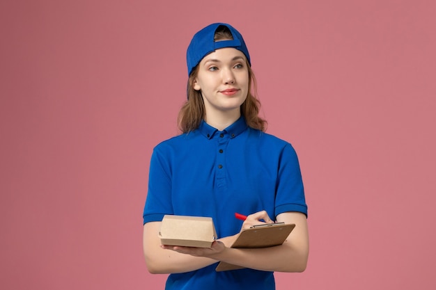 Foto gratuita mensajero femenino de vista frontal en uniforme azul y capa sosteniendo un pequeño paquete de comida de entrega y un bloc de notas escribiendo en la pared rosa, empleado del servicio de entrega