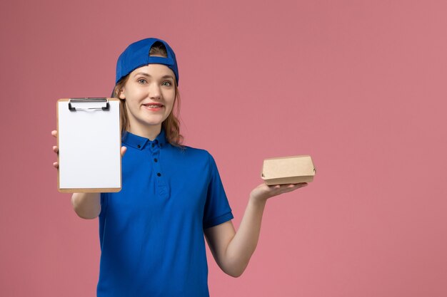 Mensajero femenino de vista frontal en uniforme azul y capa sosteniendo un pequeño paquete de comida de entrega con bloc de notas en la pared rosa, trabajo de empleado de servicio de trabajo de entrega