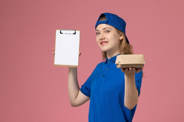 Mensajero femenino de vista frontal en uniforme azul y capa sosteniendo un pequeño paquete de comida de entrega y un bloc de notas sonriendo en la pared rosa, empleado del servicio de entrega