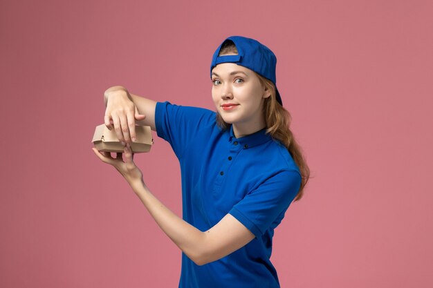 Mensajero femenino de vista frontal en uniforme azul y capa sosteniendo un pequeño paquete de comida de entrega en la empresa de servicio uniforme de trabajo de entrega de fondo rosa