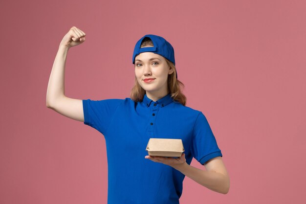 Mensajero femenino de vista frontal en uniforme azul y capa sosteniendo un pequeño paquete de comida de entrega y flexionando en la pared rosa, empresa de servicio uniforme de trabajo de entrega