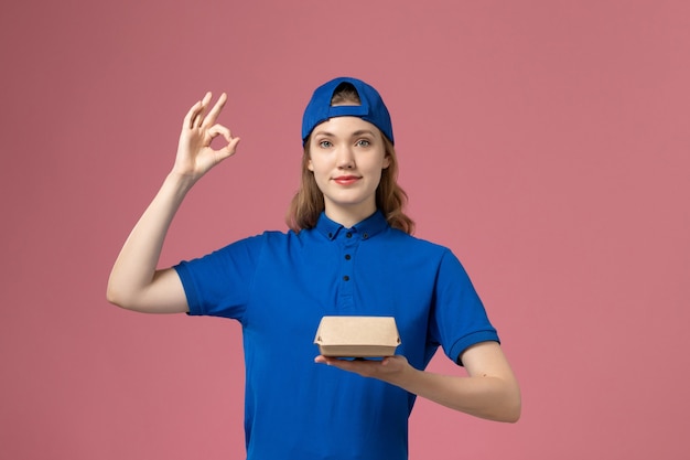 Mensajero femenino de vista frontal en uniforme azul y capa sosteniendo un pequeño paquete de comida de entrega en el fondo rosa chica de empresa de servicio uniforme de entrega