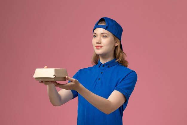 Mensajero femenino de vista frontal en uniforme azul y capa sosteniendo pequeño paquete de comida de entrega en el fondo rosa empresa de servicio uniforme de entrega trabajo trabajador niña