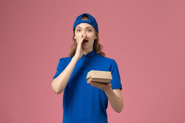 Mensajero femenino de vista frontal en uniforme azul y capa sosteniendo un pequeño paquete de comida de entrega llamando en la pared rosa, empresa de servicio de entrega uniforme