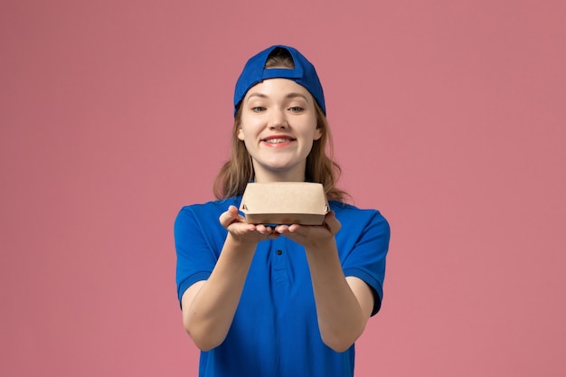 Mensajero femenino de vista frontal en uniforme azul y capa sosteniendo un pequeño paquete de comida de entrega en la pared de color rosa claro, empleado de servicio uniforme de trabajo de entrega