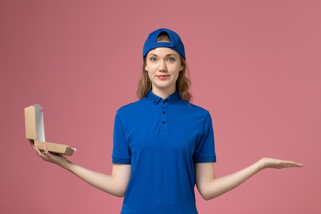 Mensajero femenino de vista frontal con uniforme azul y capa sosteniendo un pequeño paquete de comida de entrega en una pared rosa claro, trabajo de empleado de servicio uniforme de entrega