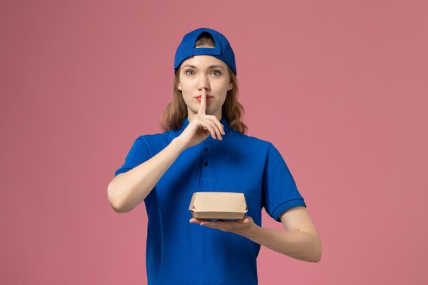 Mensajero femenino de vista frontal en uniforme azul y capa sosteniendo un pequeño paquete de comida de entrega en la pared rosa, servicio de entrega uniforme trabajo trabajador niña trabajo