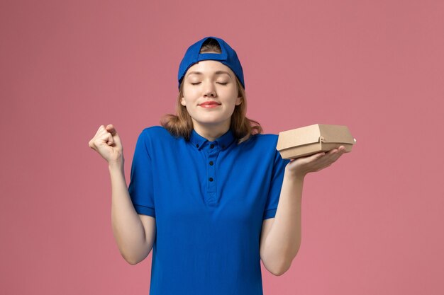 Mensajero femenino de vista frontal en uniforme azul y capa sosteniendo un pequeño paquete de comida de entrega en la pared rosa, trabajadora de la empresa de servicio uniforme de entrega de trabajo