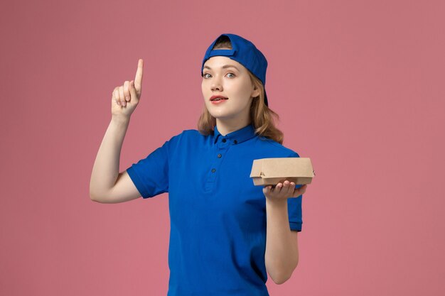 Mensajero femenino de vista frontal en uniforme azul y capa sosteniendo un pequeño paquete de comida de entrega sobre fondo rosa empresa de trabajo de servicio uniforme de entrega de trabajo