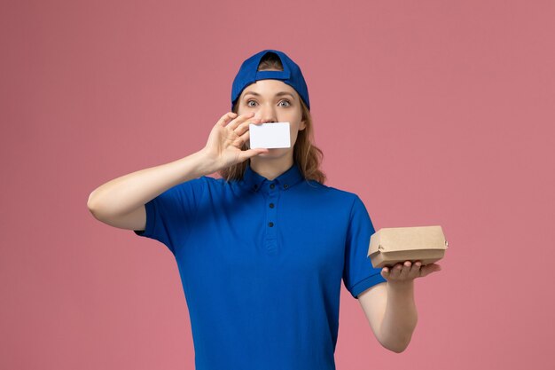 Mensajero femenino de vista frontal en uniforme azul y capa sosteniendo un pequeño paquete de comida de entrega con tarjeta en la pared rosa, empleado del servicio de entrega del trabajador