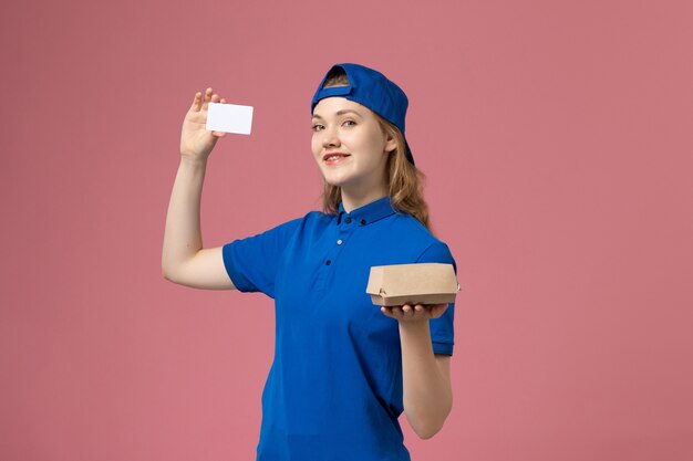 Mensajero femenino de vista frontal en uniforme azul y capa sosteniendo un pequeño paquete de comida de entrega con tarjeta en la pared rosa, empleado del servicio de trabajo de entrega