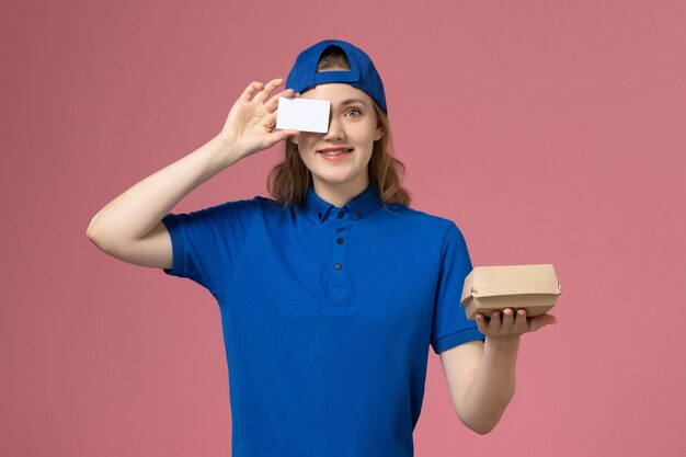 Mensajero femenino de vista frontal en uniforme azul y capa sosteniendo un pequeño paquete de comida de entrega con tarjeta en la pared rosa, trabajo de trabajo del empleado del servicio de entrega