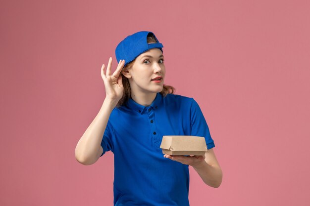 Mensajero femenino de vista frontal en uniforme azul y capa sosteniendo un pequeño paquete de comida de entrega tratando de escuchar en la pared rosa, trabajo de la empresa de servicio de entrega uniforme