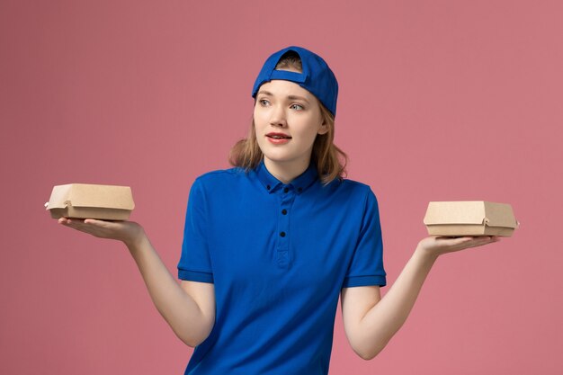 Mensajero femenino de vista frontal en uniforme azul y capa sosteniendo pequeños paquetes de comida de entrega en la pared rosa, empresa de servicio de dama de uniforme de trabajo de entrega