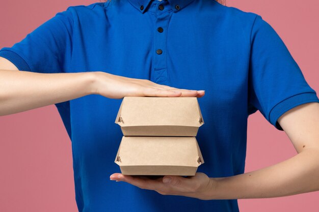 Mensajero femenino de vista frontal en uniforme azul y capa sosteniendo pequeños paquetes de comida de entrega en la pared rosa, trabajo de empresa de servicio uniforme de trabajo de entrega