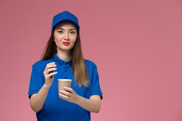 Mensajero femenino de vista frontal en uniforme azul y capa sosteniendo la taza de café de entrega abriéndola en el trabajo de la pared rosa