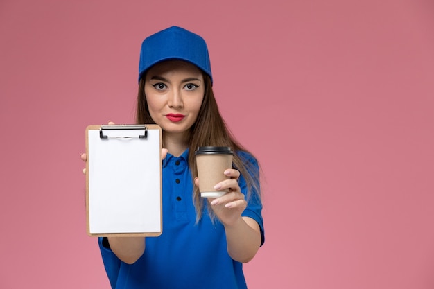 Mensajero femenino de vista frontal en uniforme azul y capa sosteniendo la taza de café de entrega y el bloc de notas en el escritorio de color rosa claro