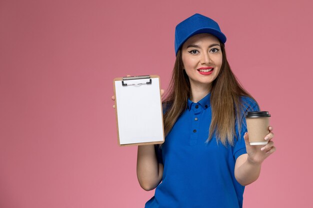 Mensajero femenino de vista frontal en uniforme azul y capa sosteniendo la taza de café de entrega y el bloc de notas en el trabajador de escritorio rosa