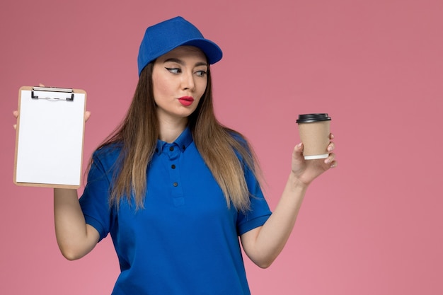 Mensajero femenino de vista frontal en uniforme azul y capa sosteniendo la taza de café de entrega y el bloc de notas en el trabajador de la pared rosa