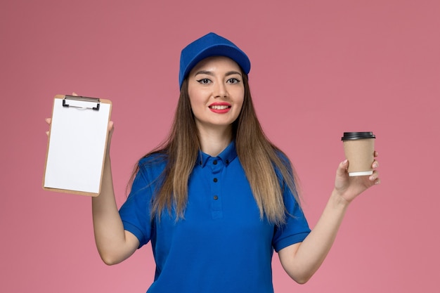 Mensajero femenino de vista frontal en uniforme azul y capa sosteniendo la taza de café de entrega y el bloc de notas en el trabajador de la pared rosa