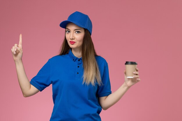 Mensajero femenino de vista frontal en uniforme azul y capa sosteniendo la taza de café de entrega en la pared rosa niña mujer