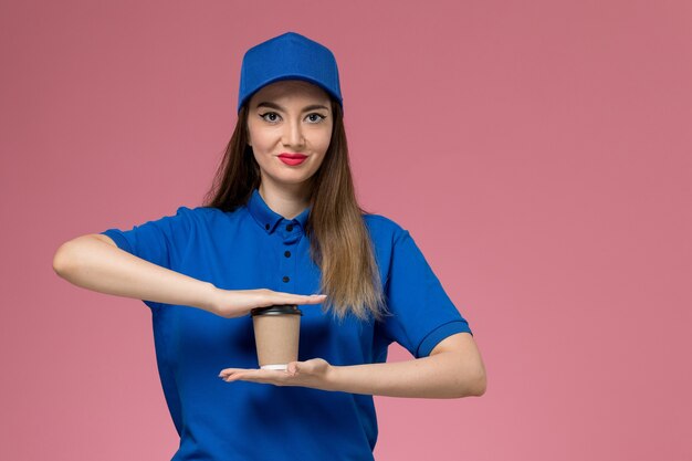 Mensajero femenino de vista frontal en uniforme azul y capa sosteniendo la taza de café de entrega en la pared rosa