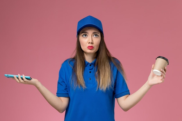 Mensajero femenino de vista frontal en uniforme azul y capa sosteniendo la taza de café de entrega usando un teléfono en la pared rosa