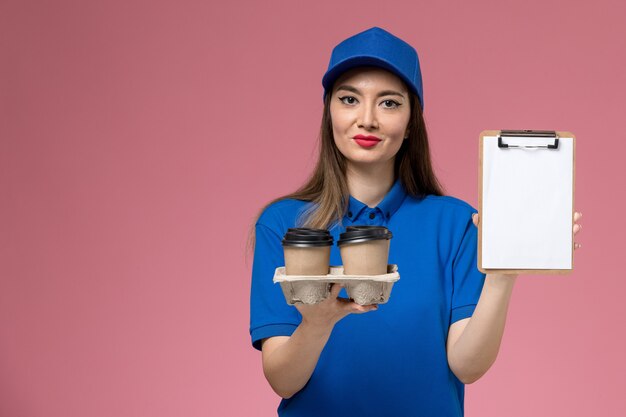 Mensajero femenino de vista frontal en uniforme azul y capa sosteniendo tazas de café con el bloc de notas en el escritorio rosa