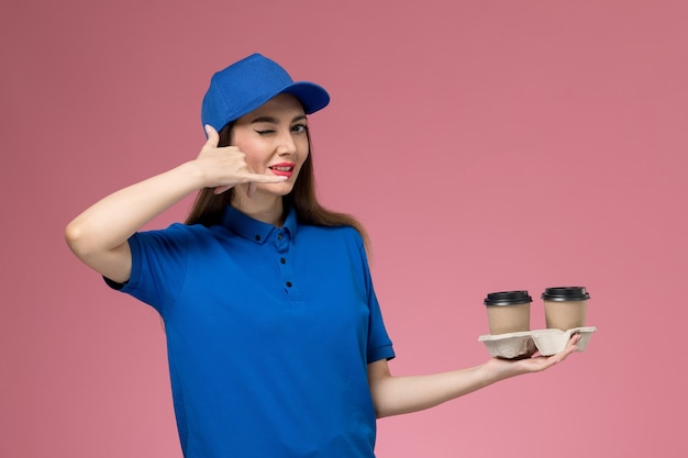 Mensajero femenino de vista frontal en uniforme azul y capa sosteniendo tazas de café de entrega guiñando un ojo en el trabajo de pared rosa