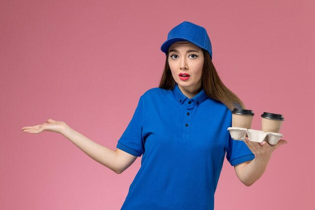 Mensajero femenino de vista frontal en uniforme azul y capa sosteniendo tazas de café de entrega marrón en la pared rosa