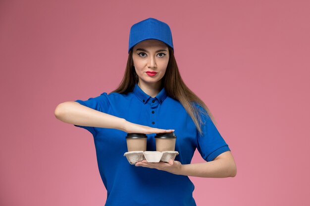 Mensajero femenino de vista frontal en uniforme azul y capa sosteniendo tazas de café de entrega marrón posando en la pared rosa