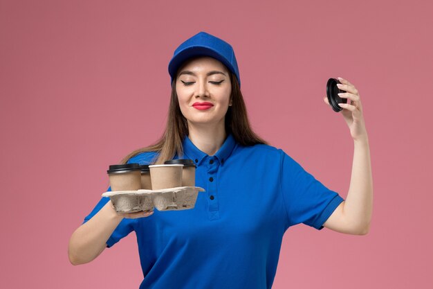 Mensajero femenino de vista frontal en uniforme azul y capa sosteniendo tazas de café de entrega y oliendo en la pared rosa