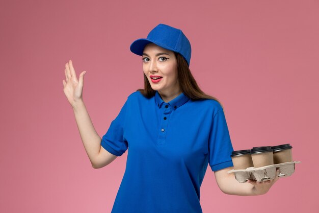 Mensajero femenino de vista frontal en uniforme azul y capa sosteniendo tazas de café de entrega posando en la pared rosa